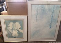 Photo showing two framed pieces side by side: watercolor irises by Robert White (left) and large blue forest print (right)