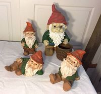 Four garden gnome figurines on white cloth with closed door in background. Three smaller gnomes with red hats, green shirts, white beards, brown shoes, and one larger gnome holding a basket.