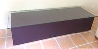 Side angle view showing the dark eggplant rectangular base and clear glass top of the coffee table/bench, positioned on a tiled floor.