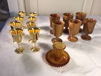 Photo showing 8 shiny gold plated stemware glasses, 6 wooden stemware glasses, and one amber-colored glass candle holder on a white cloth surface.