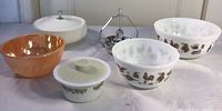 Overview of lot showing two white Pyrex bowls with brown animal designs, iridescent orange Fire King bowl, lidded Pyrex floral bowl, white lidded dish, and small floral serving dish with metal utensil.