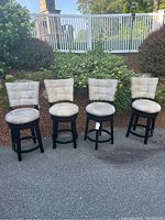 Four black swivel counter stools with beige cushioned backs and seats arranged outdoors, showing front and side views.
