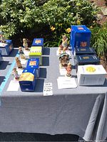 Eight Hummel figurines arranged on a table with their original boxes underneath and around them. Green shrubbery and flowers in background.