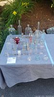 Photo showing assortment of clear glass bottles and jars arranged on a grey cloth outside, including various shapes and sizes such as bottles with narrow necks, flasks, small vials, and jars. One bottle has a red stopper.