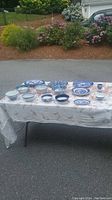 Photo of 14 assorted blue and white ceramic and porcelain serving ware pieces spread on a white tablecloth outdoors. Includes plates, bowls, and platters with various blue patterns including Blue Willow and hand painted designs.