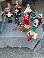 Seven Byer's Choice Carolers figurines with a red bicycle and wooden log on grey cloth in a garage setting.