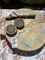 Photo of large hand drum with painted tribal symbols, two wooden shakers, and a colorful wrapped beater on patterned cloth background.