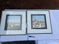 Two framed Elizabeth Mumford paintings on white tablecloth outdoors, one showing Cape Cod girls scene, the other Cape Cod boys scene with sledding children.