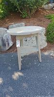 Gray round Scandinavian style end table set outdoors on pavement near rocks and plants. View of full table structure including legs and lower shelf.