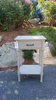 Gray wooden end table shown outdoors against rock and shrub background, displaying full front view with drawer closed.