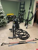 Cyclace exercise bike with battle rope and resistance bands in front, disassembled punching bag stand leaning against the wall.