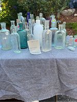 Shelf with a variety of glass bottles including clear, blue, green, and opaque white bottles arranged outdoors on grey fabric.