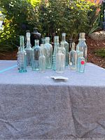 Front view of 20 assorted glass bottles arranged on a grey cloth outdoors with sunlight, showing various clear, light blue, green and amber bottles.