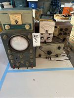 Photo shows five vintage HeathKit electronic test instruments. The largest device is a laboratory oscilloscope with multiple control knobs, an analog CRT display, and a dark-colored front panel. Other smaller meters include a reflected power meter and other tube testing instruments, all analog with dials and rotary controls.