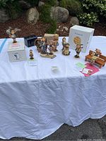 Full overview of 10 Hummel figurines arranged on table with white cloth outdoors with rocks and plants in background