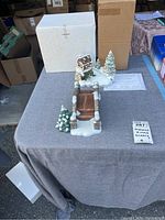 Hummel Christmas scenery figurines and decorative pieces on a grey table, includes a bridge display, Christmas tree, and a house building figurine with snow base.