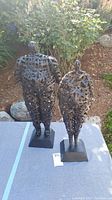 Two metal sculptures of human forms, outdoor setting on a cloth surface with plants and rocks in the background.