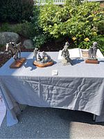 Four sculptures and open book displayed on a grey tablecloth with outdoor garden background.