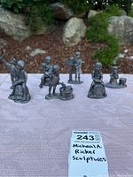 Photo of six Michael A. Ricker sculptures displayed outdoors on a white cloth with natural background. Sculptures include seated and standing figures in various poses.