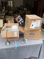 View of boxed Mitzi Hudson Valley pendant lighting components including white globe shade and old bronze finish metal canopy on a cloth-covered table in a garage setting.