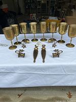 Front view of 8 brass cups standing upright with 4 candle holders and 2 bottle openers laid flat in front on a white tablecloth.