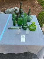 Photo of all eleven vintage glass bottles in various shades of green and clear, arranged on a gray cloth-covered surface outdoors.