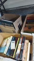 Photo showing three boxes filled with assorted books including a mix of hardback and paperback titles. Various genres represented with some cookbooks visible.