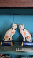 Photo of both porcelain cat figurines side by side on shelf with blue background.