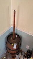 Wooden butter churn with tall dasher handle, metal lid, and barrel body. Scratches and wear visible on wood surface.