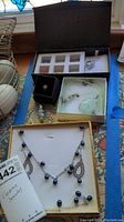 Overall view of open box with multiple jewelry items displayed including a black ring box, a green stone pendant necklace, a heart charm necklace, and a blue and silver necklace and bracelet set displayed on yellow box