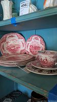 Shelf display showing five salad plates, one cracked, one teacup with saucer, and one bowl all with red transferware pattern.