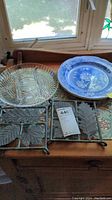 Two Spode Blue Room blue and white botanical plates, clear cut glass divided serving dish with three compartments, and two metal leaf-shaped trivets placed on a wooden surface near a window.