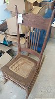 Full view of wooden rocking chair showing carved backrest, twisted spindles, cane seat, and wooden rockers. Visible wear and surface marks.
