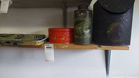 Four metal items on wood shelf: two floral burner covers, red round tin, black metal box with bottle inside