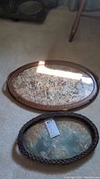 Photo of two oval wooden trays on carpet, one plain wood frame with natural material pattern, one braided wood frame with butterfly pattern.