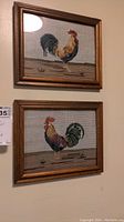 Two framed needlework rooster artworks hung vertically on a wall, showcasing their size and framing.
