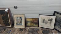 Five different framed artworks arranged leaning against a wall inside a garage space, showing variety of subjects and frame styles.