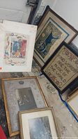 Six wall art pieces laid on the floor including framed needlework and illustrated prints in worn frames, some unframed pieces also visible.
