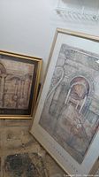 Two framed wall art prints seen leaning against a wall, one titled 'Piazza Vista' and the other 'Palace of Lilies'. Frames are gold-toned with visible water damage marks on floor and frame edges.