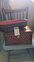 View of two items stacked on a chair: an oval metal basket with decorative floral edge and reddish interior on top; below it a rectangular wooden box with intricate geometric inlaid pattern and keyhole on front.