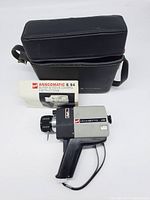 Photo showing the gaf Anscomatic S/84 Super 8 movie camera lying next to its black zippered carrying case and the instruction manual.