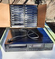 Sony Betamax player model SL-HF450 with power cord on top and box containing approximately 60 used Beta video tapes behind it.