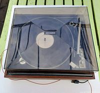 Top-view of the turntable with a vinyl record on the platter, visible tonearm, and control buttons, all enclosed under the clear dust cover.