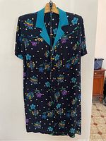 Full front view of a black silk dress with a teal collar, featuring colorful floral prints and white polka dots, short sleeves, hung on a hanger