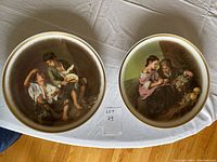 Two porcelain plates with hand-painted scenes of people, placed on a white cloth with lot number sign