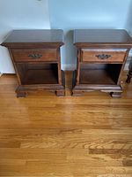 Two maple wood nightstands side by side on hardwood floor. Each has one drawer with metal handles and an open storage shelf below. Both have detailed edges and bracket feet.