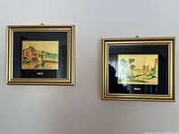 Two framed pictures on wall, each with a gold-tone frame and black mat surrounding scenic artwork on 23kt gold leaf.