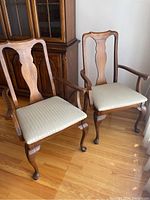Pair of wooden dining armchairs with upholstered light beige seats, curved armrests, carved backrest, cabriole front legs, and stain on wood
