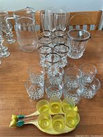 Photo of a group of clear drinking glasses with silver rims including tall and short glasses, a clear glass pitcher, an ice bucket with metal rim and tongs on a wooden table.