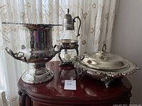Front and side view of silver ice bucket, covered serving dish, and pitcher displayed on wooden surface near curtain